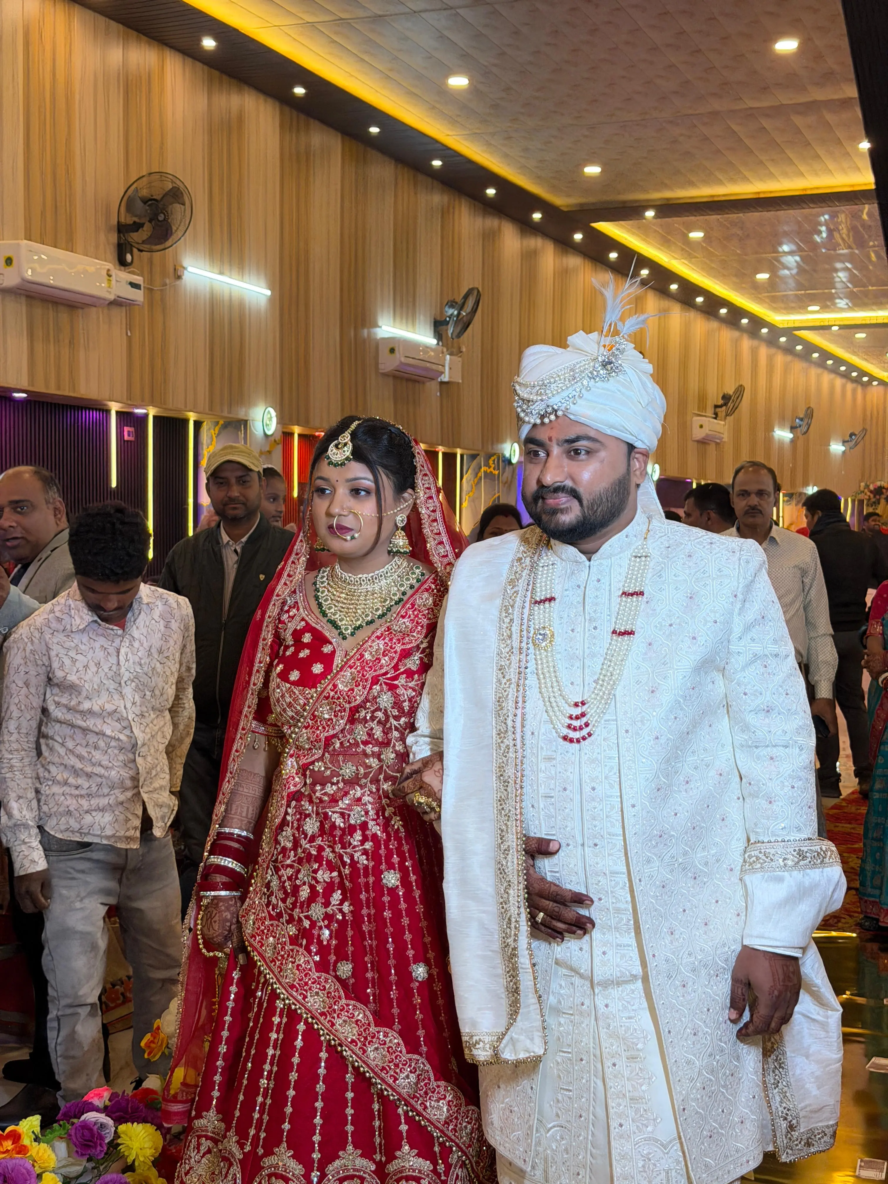 A bride in a red and gold lehenga walking arm in arm with the groom in an ivory sherwani and turban, into the wood-panelled hall